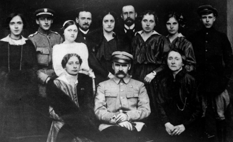 Fotografia grupowa; Józef Piłsudski (siedzi drugi z prawej) w otoczeniu rodziny i przyjaciół, między innymi poźniejszej żony Aleksandry Szczerbińskiej (Piłsudskiej) (stoi nad Naczelnikiem nieco po lewej) oraz siostry Zofii Kadenacowej (siedzi pierwsza z prawej), 1919. Źródło: NAC