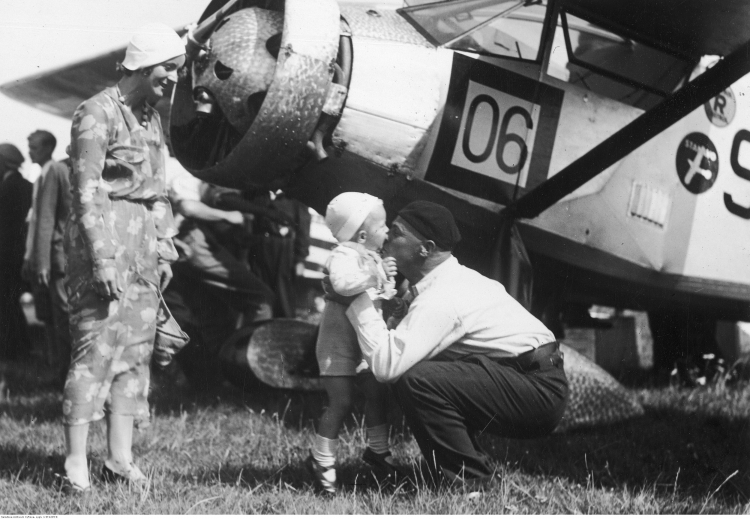 Porucznik pilot Franciszek Żwirko wita się ze swoim synkiem Heniem i żoną Agnieszką po przylocie na warszawskie lotnisko w sierpniu 1932 r.. Z tyłu widoczny samolot RWD-6. Fot. Narodowe Archiwum Cyfrowe.